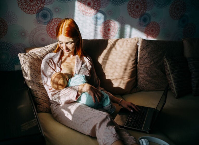 Mother breastfeeds her newborn son and works remotely with a laptop at home, sunlight from the window