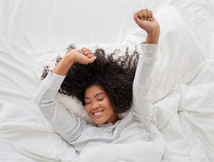 A cheerful young Hispanic woman with curly hair is waking up in a peaceful bedroom. She is stretching her arms and has a content expression on her face, top view, panorama