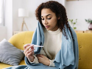 Sick young woman with cold and flu bad symptoms measuring her temperature using thermometer sitting on sofa at home