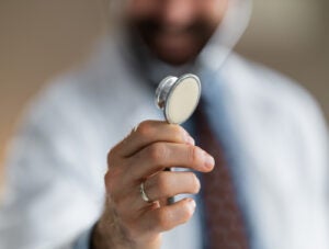 Doctor holds a stethoscope toward the camera in a close-up shot, suggesting medical examination, professional care and trust during a clinical consultation with shallow depth of field and warm tones.