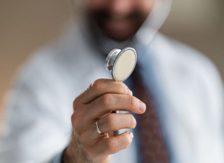Doctor holds a stethoscope toward the camera in a close-up shot, suggesting medical examination, professional care and trust during a clinical consultation with shallow depth of field and warm tones.
