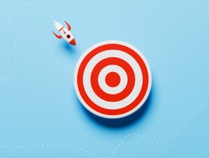 A bold conceptual illustration featuring a single red cartoon-style rocket leaving a dotted trail as it flies directly toward a central bullseye. The clean visual design highlights themes of focused execution, goal alignment, and breakthrough performance. The vivid red color emphasizes urgency, determination, and standout strategy—ideal for visuals that convey success, precision targeting, business achievement, or visionary aim in competitive or creative contexts.